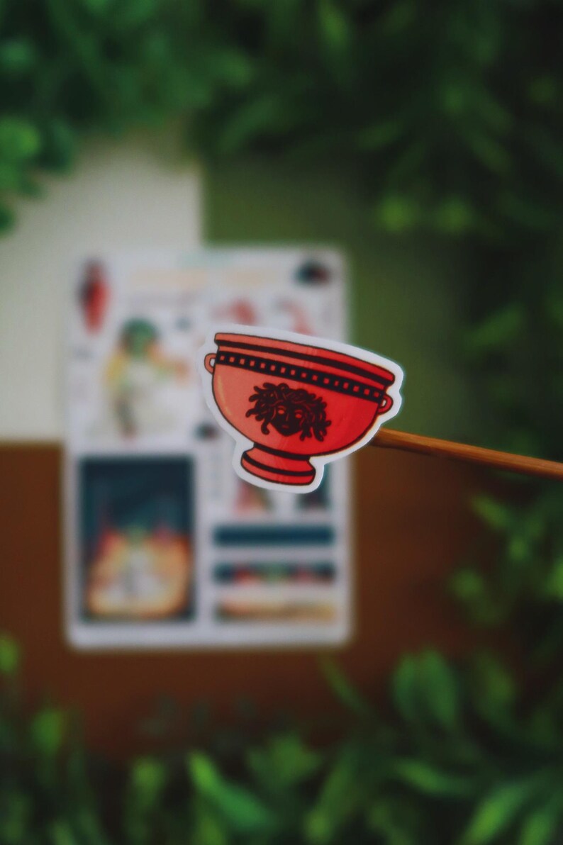 May include: A red sticker featuring a stylized bowl with a Medusa head design. The bowl has a black rim and handles. The sticker is held against a blurred background of green foliage and a white paper.
