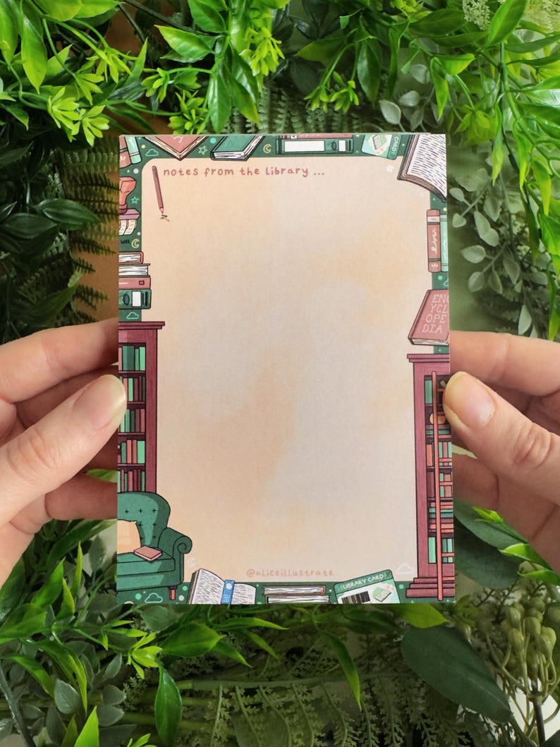 May include: A rectangular notepad with a library theme. The notepad features illustrations of bookshelves, books, and a green armchair. The text "notes from the library..." is at the top. The notepad is held by two hands.