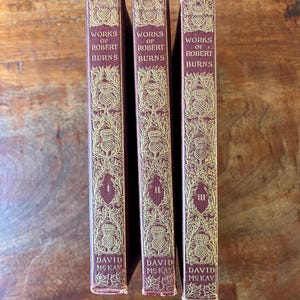 May include: Three antique books titled "Works of Robert Burns" with burgundy covers and gold decorative accents. The spines display Roman numerals I, II, and III, along with the publisher's name, David McKay.