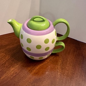 May include: A green and purple polka dot teapot with a lid. The teapot has a green handle and a purple stripe around the middle.
