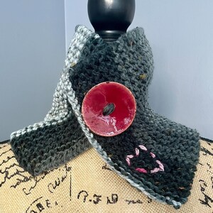 May include: A gray and white knitted scarf with a large, red, circular button. The scarf is displayed on a mannequin with a burlap-like fabric covering.