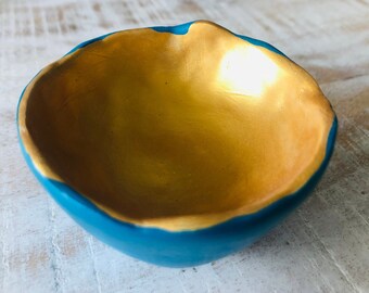 May include: A small, handmade bowl with a teal blue exterior and a gold interior. The bowl is made of clay and has a slightly uneven, organic shape.