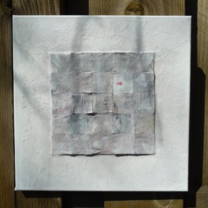 May include: A white canvas with a textured surface. A square collage of gray and white paper squares is centered on the canvas. A small pink flower is visible in the center of the collage.