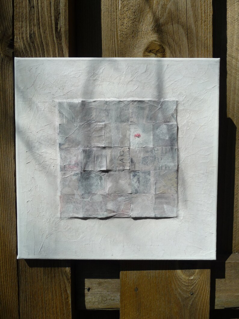 May include: A white canvas with a textured surface. A square collage of gray and white paper squares is centered on the canvas. A small pink flower is visible in the center of the collage.