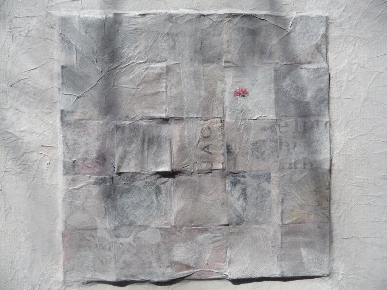 May include: A gray and white abstract art piece made of layered paper squares. The paper has a textured, wrinkled appearance. A small red dot is visible in the center of the piece.