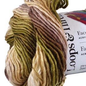 Puede incluir: Una madeja de hilo jaspeado que presenta una mezcla de fibras de color verde oliva, bronceado, marr&oacute;n y crema con un ligero brillo. Una etiqueta de papel blanco envuelve el hilo, mostrando el texto Loops & Threads, Facets y 256 metros.