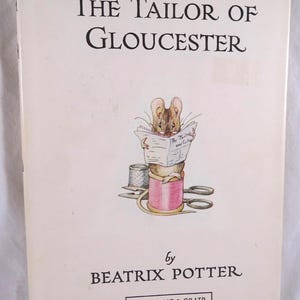 May include: A hardcover book titled The Tailor of Gloucester by Beatrix Potter. The white cover features an illustration of a mouse wearing glasses, reading a newspaper while sitting on a pink spool of thread, with scissors and a thimble nearby.