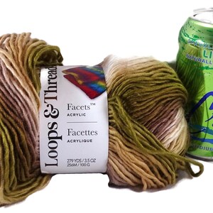 Puede incluir: Una madeja de hilo acr&iacute;lico Loops & Threads Facets con una paleta de colores variados en verde oliva, beige y crema, con una etiqueta de papel que indica 256m y 100g, colocada junto a una lata de agua con gas sabor lima LaCroix de 355ml.