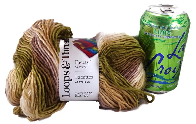 Puede incluir: Una madeja de hilo acr&iacute;lico Loops & Threads Facets con una paleta de colores variados en verde oliva, beige y crema, con una etiqueta de papel que indica 256m y 100g, colocada junto a una lata de agua con gas sabor lima LaCroix de 355ml.