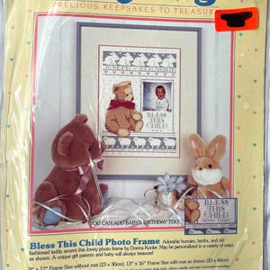 May include: BabyHugs photo frame kit with a cross-stitch design. The kit includes a framed picture with a teddy bear, a baby photo, and the text "Bless This Child." The frame size is 9" x 12" without a mat.