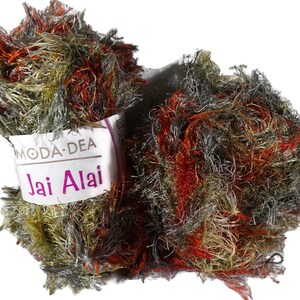 May include: A close-up of two balls of Moda Dea Jai Alai yarn. The yarn is a mix of colors, including red, gray, gold, and green. The label is white with the words "Moda Dea" and "Jai Alai" in purple.