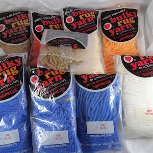 May include: Multiple packages of Red Heart Bulky Rug Yarn by Coats & Clark, each containing 320 pieces of 100% Orlon acrylic fiber. The yarn comes in various colors including blue, white, orange, and tan, with labels indicating matched dye lots.