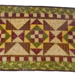 May include: Rectangular quilt with a patchwork design. The quilt features a central pattern of squares and triangles in shades of brown, green, yellow, and burgundy. A green floral border frames the design.