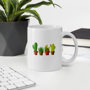 May include: White ceramic mug featuring a design of four potted cacti in a line. The cacti are green and brown with pink flowers.