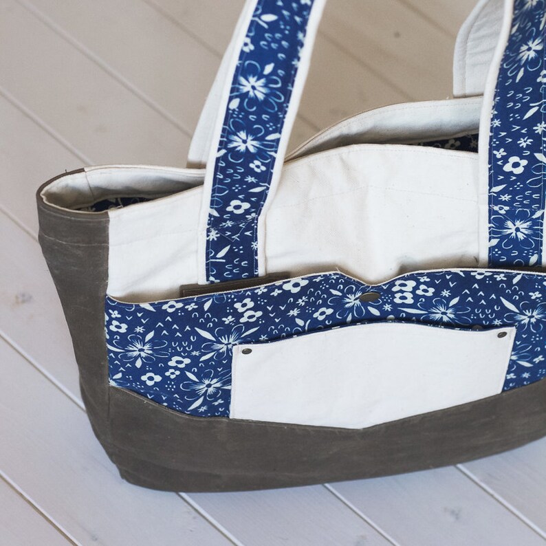 May include: A canvas tote bag with a dark gray base and white interior. The bag features blue floral patterned accents on the pockets and straps. The straps are white with blue floral designs. The bag has multiple pockets.