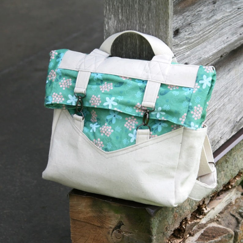 K&ouml;nnte beinhalten: Ein wei&szlig;er Canvas-Rucksack mit einer gr&uuml;nen, floralen Klappe und schwarzen Metallschnallen. Der Rucksack hat einen Tragegriff oben und verstellbare Tr&auml;ger.