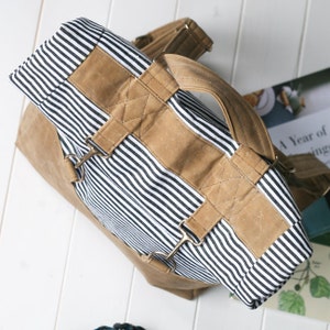 K&ouml;nnte beinhalten: Ein gestreifter Canvas-Rucksack mit braunen Lederriemen und Schnallen. Der Rucksack hat einen Tragegriff oben und verstellbare Tr&auml;ger.