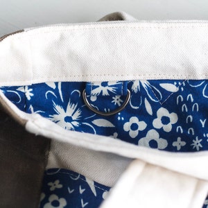 May include: Close-up of a canvas tote bag with a white top and a blue floral patterned interior. The bag has a silver metal ring attached to the interior. The exterior is a neutral brown color.