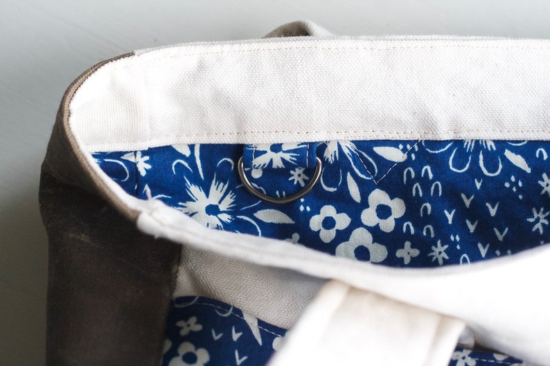 May include: Close-up of a canvas tote bag with a white top and a blue floral patterned interior. The bag has a silver metal ring attached to the interior. The exterior is a neutral brown color.