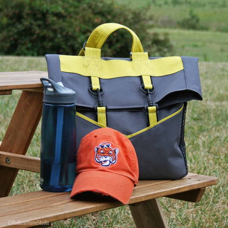 K&ouml;nnte beinhalten: Eine graue und gelbe Tragetasche mit einem Netzfach und einer leuchtend orangefarbenen Baseballkappe mit einem Tiger-Maskottchen-Logo. Die Tasche hat zwei verstellbare Tr&auml;ger und einen Griff. Die Kappe liegt auf einem h&ouml;lzernen Picknicktisch.