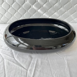 May include: A glossy black oval-shaped ceramic bowl. The bowl has a smooth, reflective surface and a wide, shallow design. The interior is also black, and the bowl sits on a white surface. The background is a white quilted pattern.