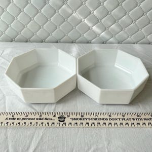 May include: Two white, octagonal bowls are displayed side-by-side. The bowls have a simple, modern design. A wooden ruler with text is in the foreground. The text on the ruler reads "A rule to prevent wild fires..." and "SMOKEY'S FRIENDS DON'T PLAY WITH MAT".