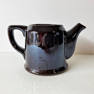 May include: A dark brown ceramic teapot with a curved handle and a spout. The teapot has a glossy finish and a rounded base. The handle is a curved shape, and the spout extends from the body. The teapot is designed for brewing tea.
