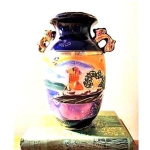 May include: A vintage ceramic vase with a lid, featuring a colorful landscape design. The vase has a dark blue top, orange and yellow midsection, and a purple base. It has two ornate handles and sits on a green book.