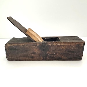 May include: A vintage wooden hand plane with a dark, weathered appearance. The plane features a rectangular wooden body with a metal blade and a wooden wedge. The blade is angled upwards, and the tool shows signs of age and use.