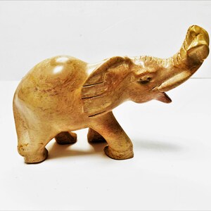 May include: A hand-carved stone elephant sculpture with a light brown color and a textured surface. The elephant's trunk is raised in the air.