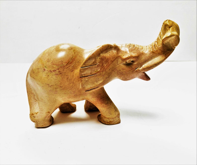 May include: A hand-carved stone elephant sculpture with a light brown color and a textured surface. The elephant's trunk is raised in the air.