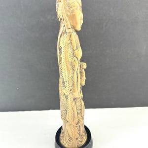 May include: A side view of a detailed, golden-colored figurine of a person in traditional attire. The figurine is mounted on a black, circular base. The figure's clothing and headdress feature intricate carvings.