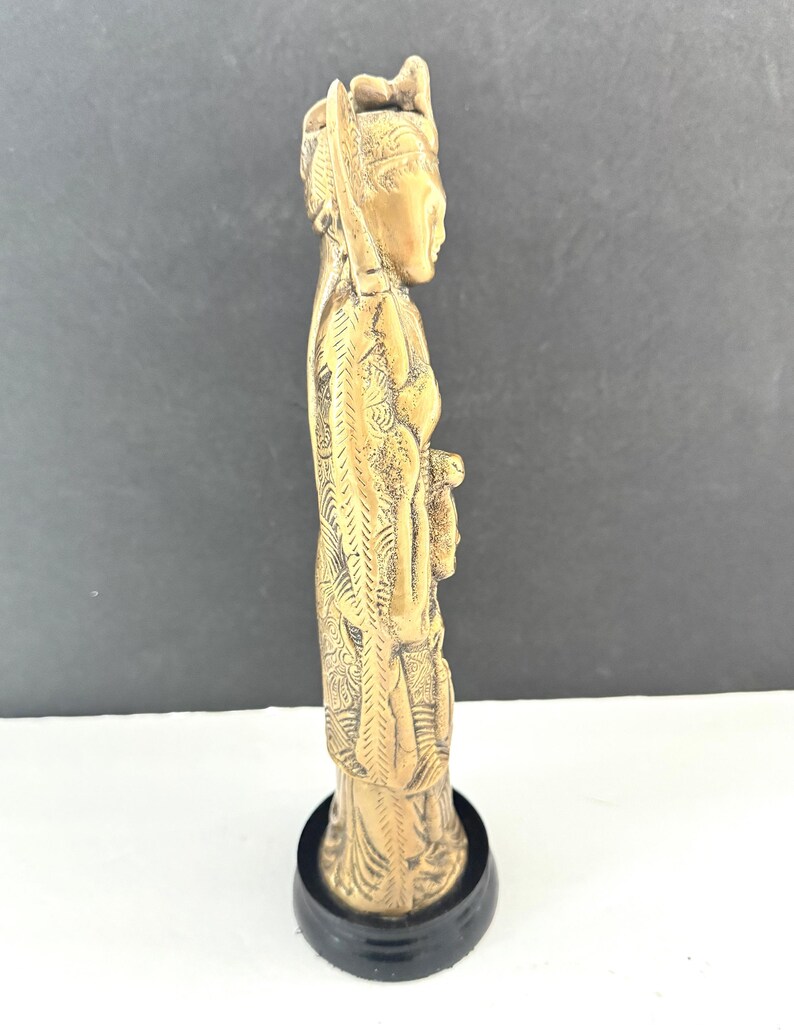 May include: A side view of a detailed, golden-colored figurine of a person in traditional attire. The figurine is mounted on a black, circular base. The figure's clothing and headdress feature intricate carvings.
