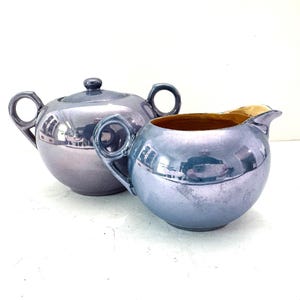 May include: A vintage tea set featuring a sugar bowl with a lid and a creamer. Both pieces have a metallic, iridescent finish in shades of blue and silver. The creamer has a gold interior.