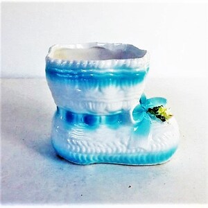May include: A white ceramic boot-shaped planter featuring a glossy finish with bright blue painted accents along the top rim and base. The front of the boot is decorated with a moulded blue bow and a small cluster of yellow and green floral details.
