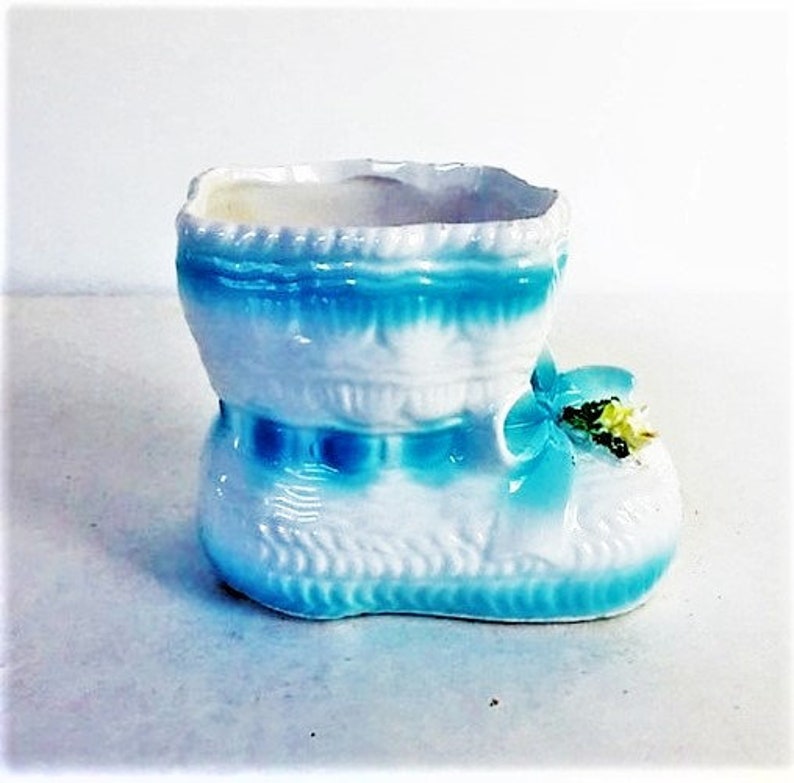 May include: A white ceramic boot-shaped planter featuring a glossy finish with bright blue painted accents along the top rim and base. The front of the boot is decorated with a moulded blue bow and a small cluster of yellow and green floral details.