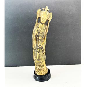 May include: A gold-colored figurine of a standing figure with ornate details, including a headdress and flowing robes. The figurine is mounted on a black circular base. The figure is holding objects in its hands. The background is a gradient of black and white.