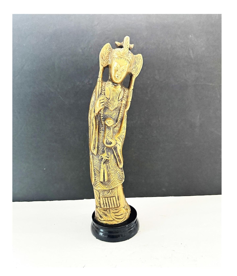 May include: A gold-colored figurine of a standing figure with ornate details, including a headdress and flowing robes. The figurine is mounted on a black circular base. The figure is holding objects in its hands. The background is a gradient of black and white.