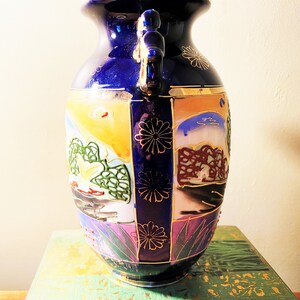 May include: A cobalt blue ceramic vase with two handles and a wide mouth. The vase features hand-painted landscape scenes in shades of yellow, orange, green, and purple, accented with gold trim and floral designs. A decorative piece.