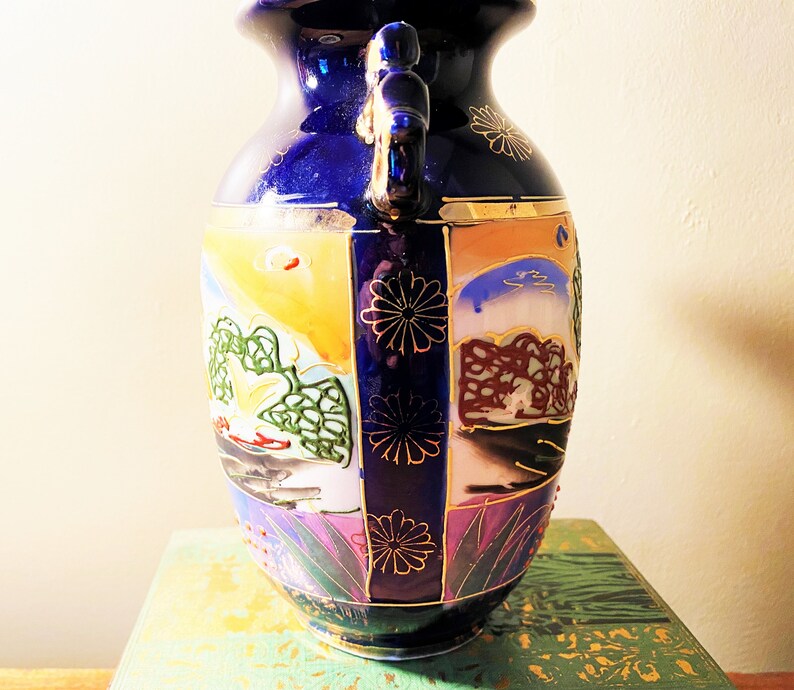 May include: A cobalt blue ceramic vase with two handles and a wide mouth. The vase features hand-painted landscape scenes in shades of yellow, orange, green, and purple, accented with gold trim and floral designs. A decorative piece.