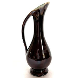 May include: A dark brown ceramic pitcher with a flared spout and a curved handle. The pitcher has a glossy finish and a slight green glaze.