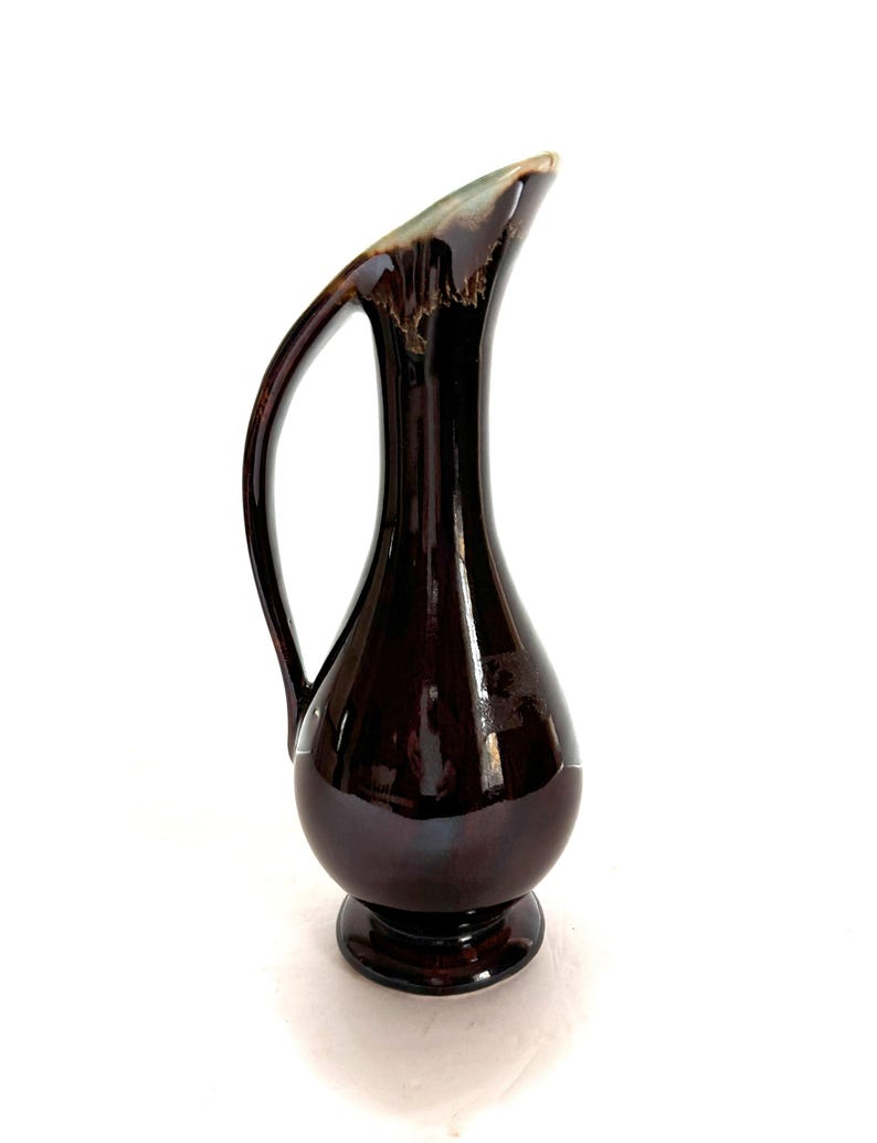 May include: A dark brown ceramic pitcher with a flared spout and a curved handle. The pitcher has a glossy finish and a slight green glaze.