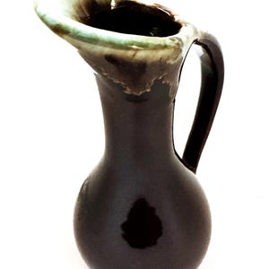 May include: A small brown ceramic pitcher with a green glazed rim and a handle. The pitcher has a rounded body and a narrow neck.
