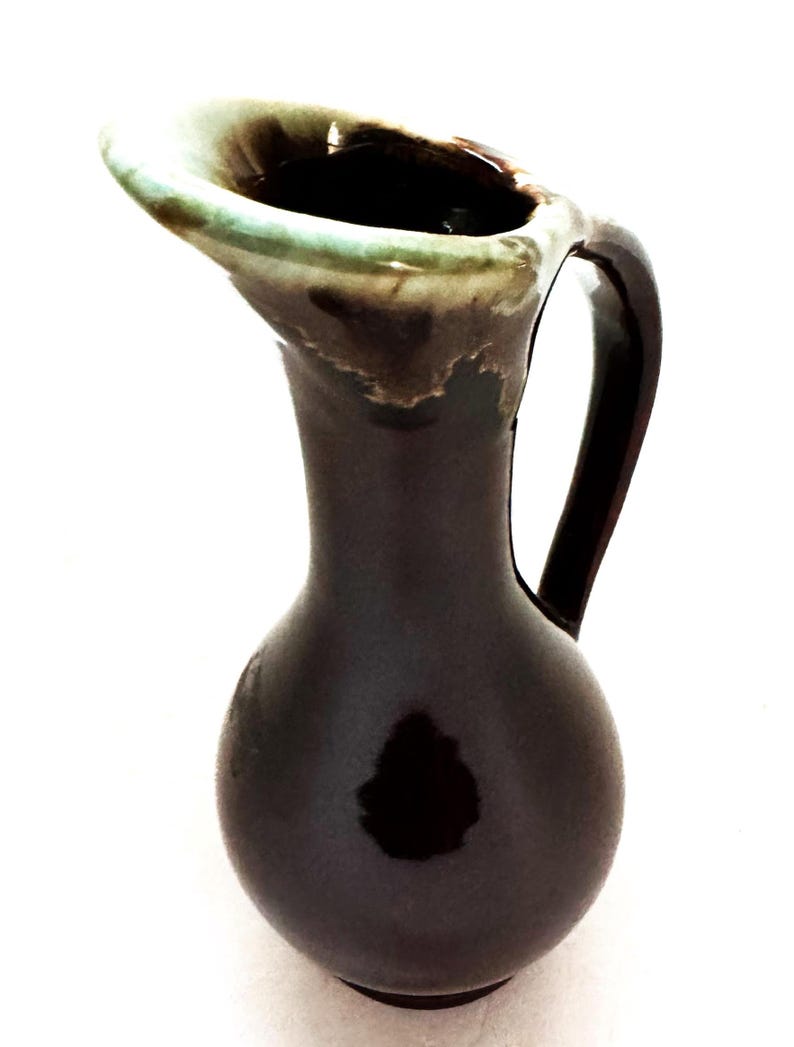 May include: A small brown ceramic pitcher with a green glazed rim and a handle. The pitcher has a rounded body and a narrow neck.
