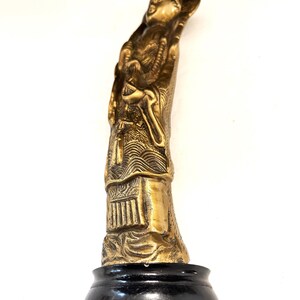 May include: A gold-colored statue of a robed figure with a headdress, standing on a black base. The statue has intricate details, including facial features, clothing folds, and decorative elements. The figure appears to be holding an object. The statue is set against a white background.