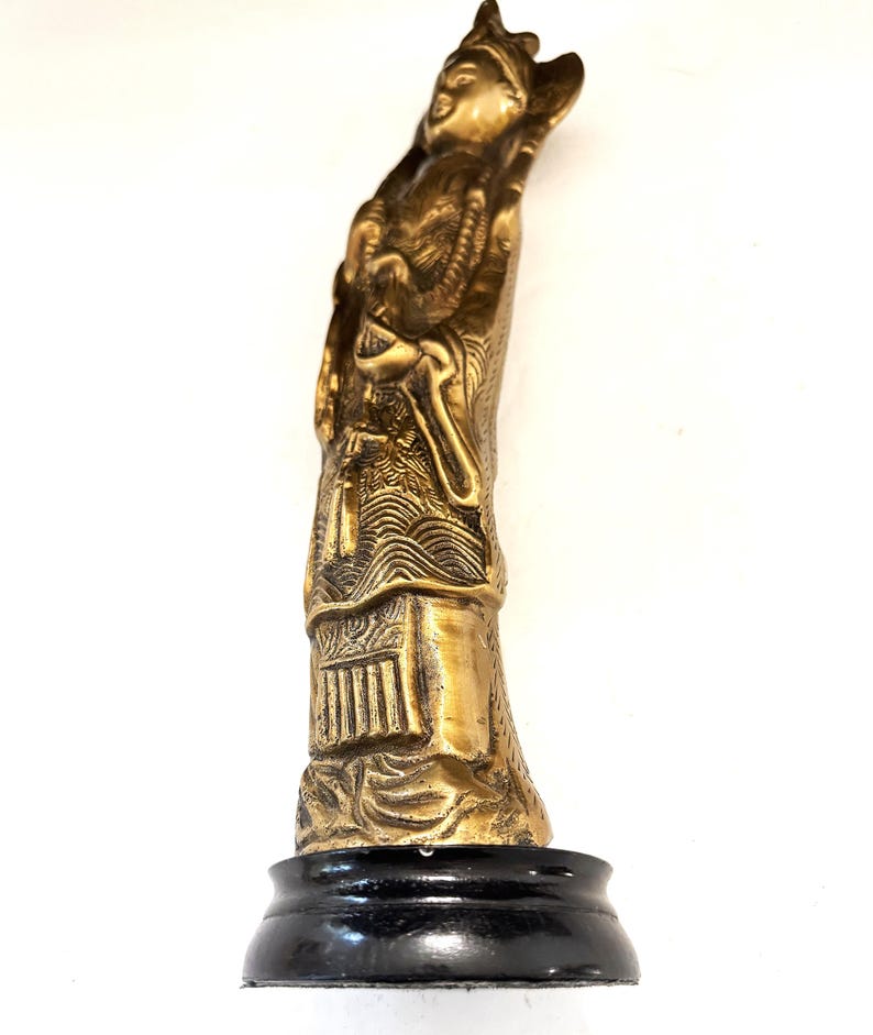 May include: A gold-colored statue of a robed figure with a headdress, standing on a black base. The statue has intricate details, including facial features, clothing folds, and decorative elements. The figure appears to be holding an object. The statue is set against a white background.