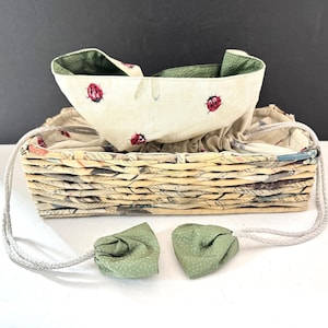 May include: A beige fabric basket with a matching drawstring bag featuring red ladybug designs. The basket is made of woven paper and has a green interior. Two green fabric accents with drawstring closures are attached.