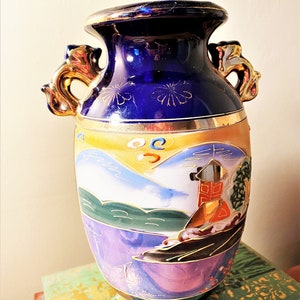 May include: A vintage ceramic vase with a dark blue top and gold accents. The vase features a painted landscape scene with mountains, water, and a building. It has two ornate gold handles and is approximately 10 inches tall.