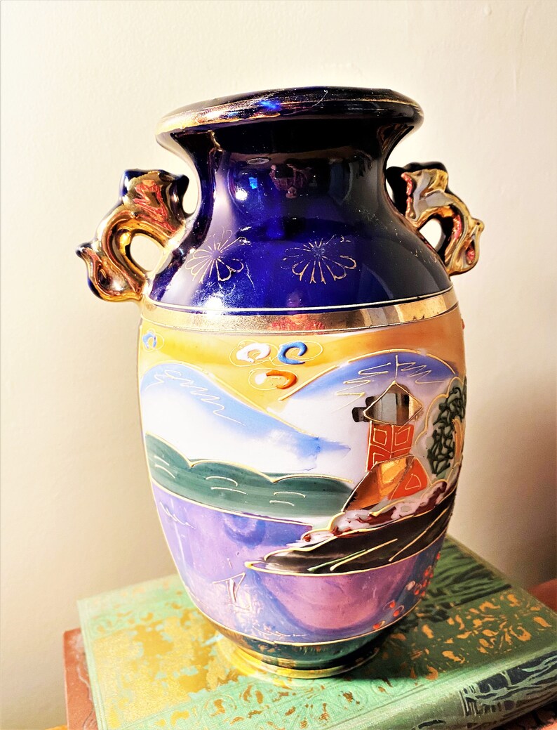 May include: A vintage ceramic vase with a dark blue top and gold accents. The vase features a painted landscape scene with mountains, water, and a building. It has two ornate gold handles and is approximately 10 inches tall.