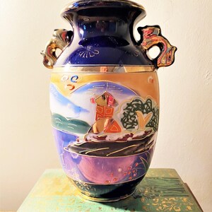 May include: A vintage, hand-painted ceramic vase with a dark blue top and gold accents. The body features a landscape scene with a red building, mountains, and trees. The vase has two ornate handles and sits on a green book.